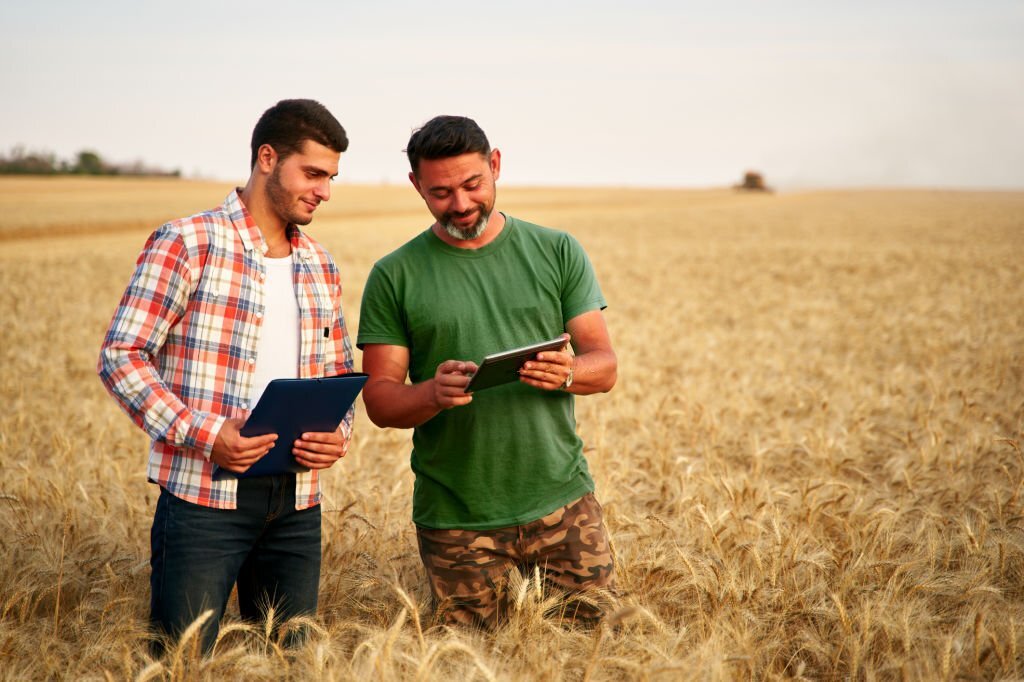 Two farmers stand in wheat stubble field, discuss harvest, crops. Senior agronomist with touch tablet pc teaches young coworker. Innovative tech. Precision farming with online data management software